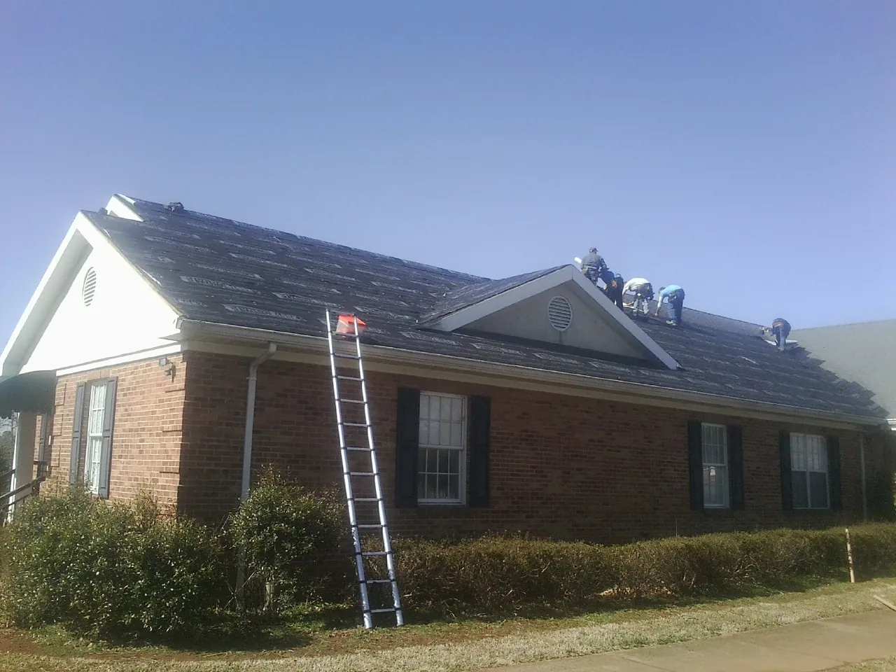Crew tearing shingles off a brick ranch roof with a ladder set — Metal Roof Repair work by our crew in Bruington
