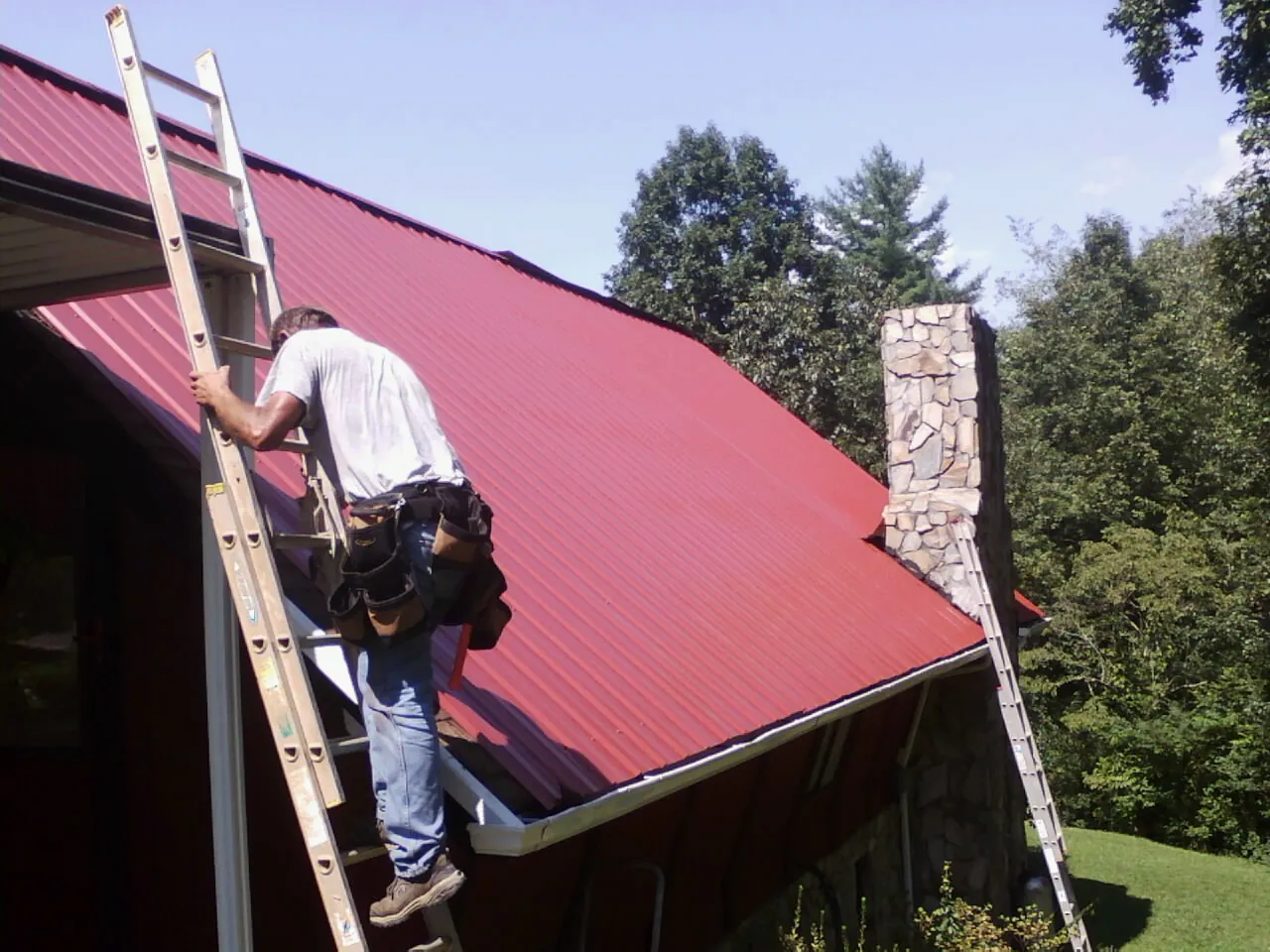 Roofer on a ladder beside a red metal roof and stone chimney — Soffit & Fascia Repair by Roofing Brothers in Bruington